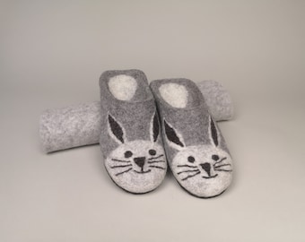 Op de afbeelding: Een paar grijze vilten konijnenslippers met witte binnenkant en zwart geborduurde gezichtskenmerken. De slippers staan naast een opgerold vilt item, alles op een neutrale achtergrond.