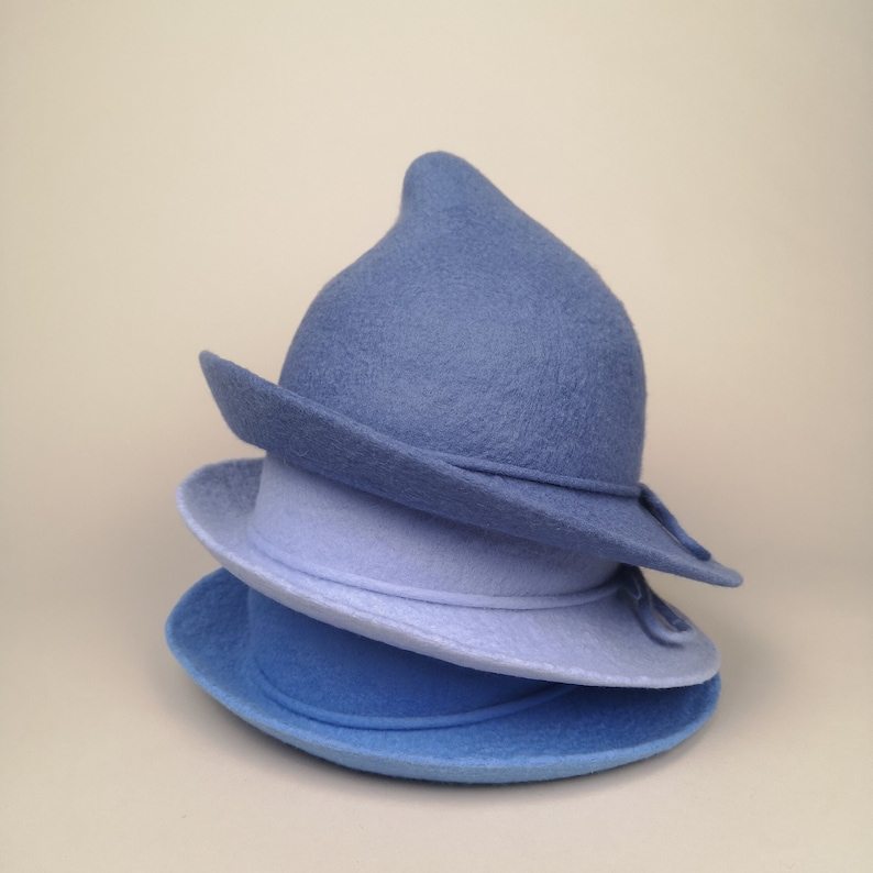 May include: Three felt hats stacked on top of each other. The top hat is a pointed, blue felt hat. The middle hat is a light blue felt hat. The bottom hat is a darker blue felt hat.