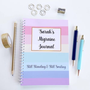 May include: A pink, blue, and white striped spiral bound journal with the text "Sarah's Migraine Journal" and the phrase "Still Standing & Still Smiling" on the cover. The journal is surrounded by gold paper clips, a pencil, and two pens.