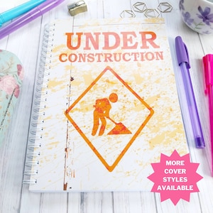 May include: A spiral bound notebook with a white background and orange and yellow splatter paint. The cover features a black and orange construction sign with a worker and the text "UNDER CONSTRUCTION". The text "MORE COVER STYLES AVAILABLE" is in a pink starburst.
