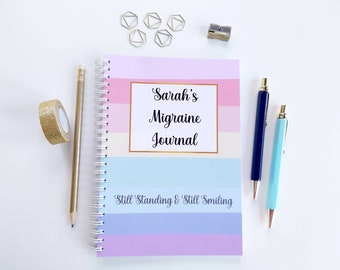 May include: A pink, blue, and white striped spiral bound journal with the text "Sarah's Migraine Journal" and the phrase "Still Standing & Still Smiling" on the cover. The journal is surrounded by gold paper clips, a pencil, and two pens.