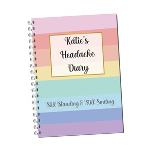 May include: A spiral bound notebook with a rainbow striped cover. The cover has a gold frame with the text "Katie's Headache Diary" and the phrase "Still Standing & Still Smiling" at the bottom.