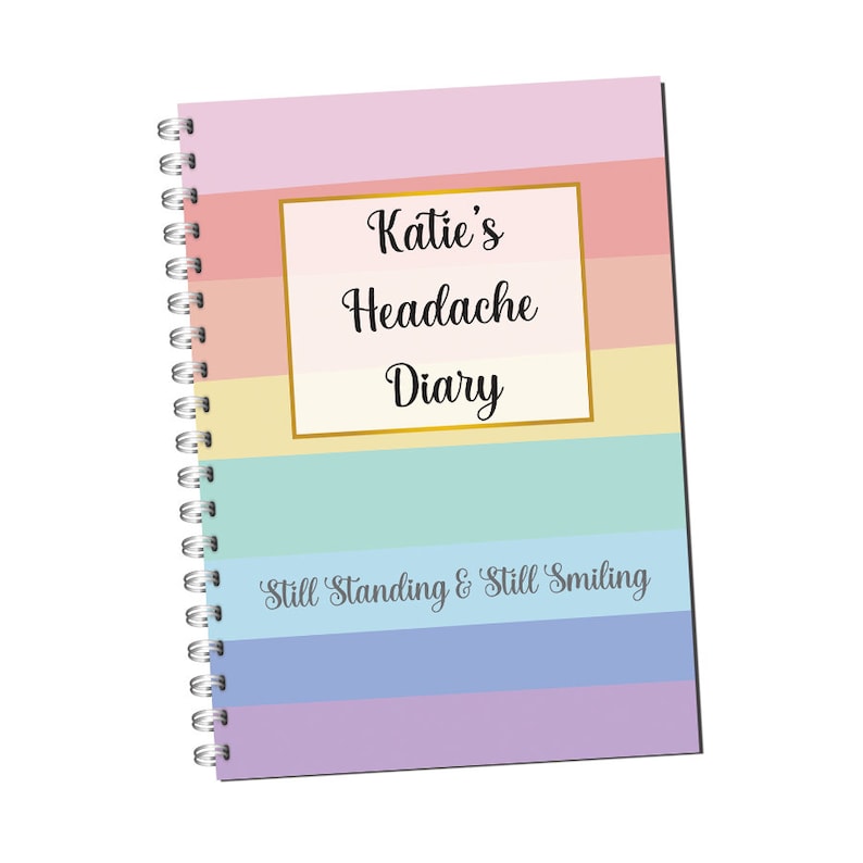 May include: A spiral bound notebook with a rainbow striped cover. The cover has a gold frame with the text "Katie's Headache Diary" and the phrase "Still Standing & Still Smiling" at the bottom.