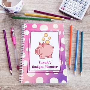 May include: A pink spiral bound notebook with a white polka dot pattern and a pink piggy bank illustration. The piggy bank has gold coins falling into it. The text "Sarah's Budget Planner" is printed on the cover.