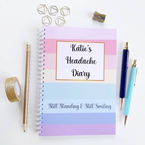 May include: A pink, blue, and white striped journal with the text "Katie's Headache Diary" and the phrase "Still Standing & Still Smiling". The journal is closed and has a gold spiral binding. There are gold paper clips, a pencil, two pens, and a pencil sharpener on a white surface.