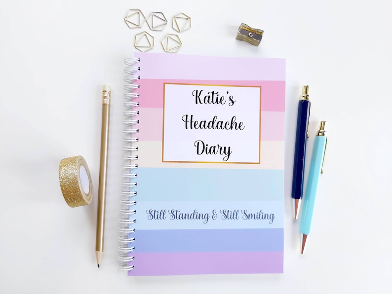 May include: A pink, blue, and white striped journal with the text "Katie's Headache Diary" and the phrase "Still Standing & Still Smiling". The journal is closed and has a gold spiral binding. There are gold paper clips, a pencil, two pens, and a pencil sharpener on a white surface.