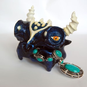 Puede incluir: Una figurita de cerámica azul de una criatura con cuernos blancos y joyas de piedras preciosas turquesa. La criatura tiene un ojo grande de color naranja y una superficie detallada y texturizada.
