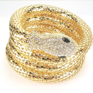 Könnte beinhalten: Ein goldfarbenes Schlangenarmband mit gewickeltem Design. Der Schlangenkopf ist mit klaren Kristallen und einem schwarzen Auge verziert. Das Armband besteht aus einem strukturierten, metallischen Material.