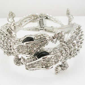 Könnte beinhalten: Ein silbernes Alligator-Armband mit schwarzen Akzenten und klaren Strasssteinen. Das Armband ist mit einem Scharnier versehen und hat einen Federschnappverschluss.