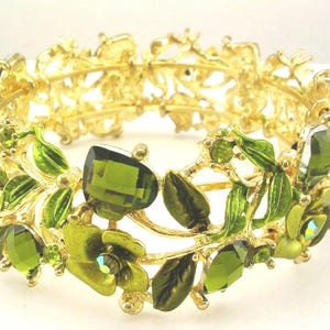 May include: Gold-coloured bracelet with a floral design. The bracelet features green gemstones and green enamel leaves and flowers. The bracelet has a hinged closure. The bracelet is approximately 5 cm in diameter.
