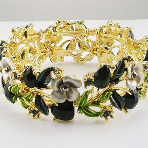 Könnte beinhalten: Goldfarbenes Armband mit floralem Design, schwarzen und grauen Edelsteinen, grünen Emailleblättern und weißen Blüten. Das Armband hat ein aufwendiges, offenes Design.
