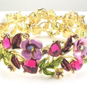 May include: A gold bracelet with a floral design. It features purple and fuchsia gemstones and flowers, with green enamel leaves. This elegant bracelet is a colourful accessory, perfect for special occasions.