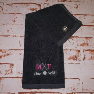 Può includere: Un asciugamano da golf grigio scuro con un design di mazza da golf e palla bianco. L'asciugamano ha un anello per appenderlo e le iniziali "M P" ricamate in rosa.