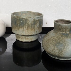 Könnte beinhalten: Drei Keramik-Töpfe in Grün- und Brauntönen. Der Topf links ist ein runder, weißer Topf mit einem Holzstiel. Der mittlere Topf ist ein höherer, zylindrischer Topf mit einer breiten Basis. Der Topf rechts ist ein runder Topf mit einem Henkel.