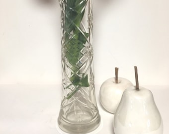 May include: A clear glass vase with a cut glass design. The vase is filled with green leaves and a single red and yellow tulip. Two white ceramic pears are in the foreground.