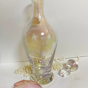 May include: A clear iridescent glass decanter with a round base and a stopper. The decanter is sitting on a white surface with a string of pearls and a pink tulip.