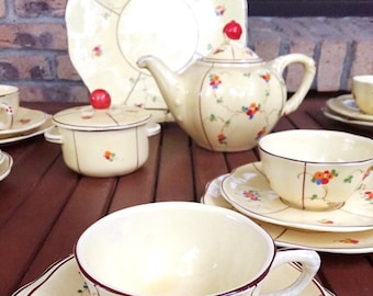 Op de afbeelding: Een vintage theeservies met een crèmekleurige basis en bloemenmotieven in rood, oranje en groen. De set bevat een theepot, roomkan, suikerpot en twee theekopjes met schoteltjes.