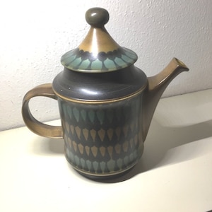 May include: A brown and green ceramic teapot with a geometric pattern. The teapot has a lid with a pointed top.
