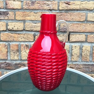 Op de afbeelding: Een rode glazen kan met een touwachtig ontwerp. De kan heeft een heldere glazen handvat en een brede opening.