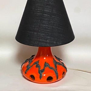 Op de afbeelding: Een kleine tafellamp met een zwarte lampenkap en een feloranje voet. De voet heeft een textuurontwerp met donkergrijze accenten en cirkelvormige uitsparingen. De lamp heeft een wit snoer.