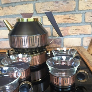 May include: A black and gold teapot with a copper and gold geometric design sits on a matching stand with four matching copper and gold teacups with handles.