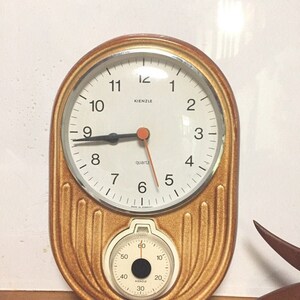 Puede incluir: Un reloj ovalado de color marrón y dorado con una esfera blanca y agujas negras. El reloj tiene un movimiento de cuarzo y un pequeño temporizador en la parte inferior. El reloj está hecho en Alemania.