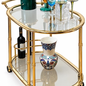 May include: An oval gold-toned metal bar cart with two glass shelves on wheels. The top shelf holds a green ice bucket, a blue glass, a crystal decanter, and two stemmed glasses. The bottom shelf holds a wine bottle and a floral ceramic vase.