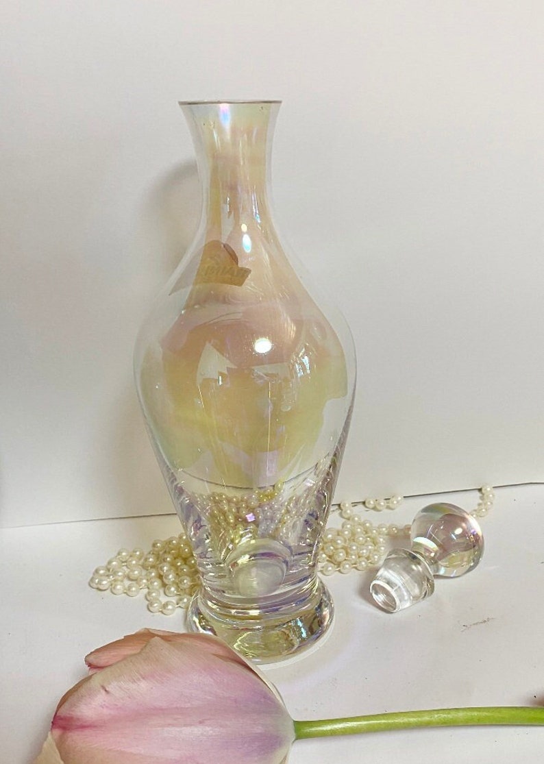 May include: A clear iridescent glass decanter with a round base and a stopper. The decanter is sitting on a white surface with a string of pearls and a pink tulip.