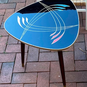 Puede incluir: Una mesa auxiliar de estilo moderno de mediados de siglo con una tapa triangular de vidrio. La tapa de vidrio presenta un diseño geométrico en tonos de azul, rosa y amarillo. La mesa tiene tres patas de madera oscura con detalles dorados.