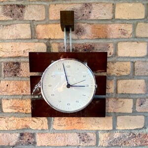 Può includere: Un orologio argentato con quadrante bianco e lancette nere è appeso a un muro di mattoni. L'orologio è montato su una cornice in legno marrone scuro.