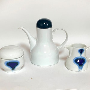 Puede incluir: Una tetera de cerámica blanca con tapa negra, una jarra de crema de cerámica blanca y un azucarero de cerámica blanca. Las tres piezas tienen un diseño abstracto azul y morado.