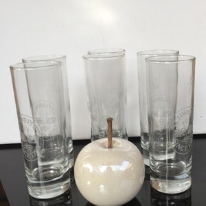 May include: Six clear glass shot glasses with a white logo that says "Schweppes Since 1783" on the front. A white ceramic apple sits in the center of the glasses.