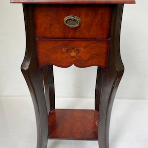 Può includere: Tavolino laterale in legno antico con un cassetto e un ripiano inferiore. Il tavolo presenta una struttura marrone scuro e un frontale del cassetto marrone rossiccio con una maniglia decorativa in ottone. Il cassetto ha un design intarsiato. Il tavolo è alto 71 cm.