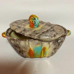 以下が含まれることがあります： 蓋付きのヴィンテージセラミック製サービングディッシュ。 皿は、オレンジ、青、黄色のアクセントが付いた、まだらの茶色とクリーム色の外観をしています。 蓋にはカラフルな渦巻きハンドルが付いています。 この皿は、食べ物を提供するように設計されています。