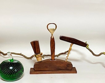 Op de afbeelding: Een houten wijnset met een flesopener, kurkentrekker en flesstopper. De tools hebben messing accenten en staan in een donkerhouten houder. Een groene glazen appel staat op de achtergrond.