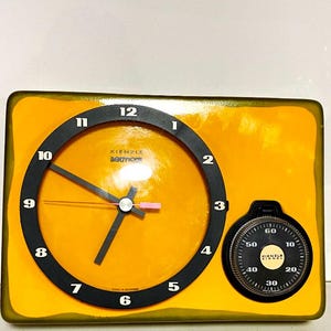 Peut inclure: Horloge vintage Kienzle BOOTICO avec un cadran jaune vif et des détails noirs. L'horloge comprend un cadran rond et un petit cadran de minuterie. Le cadran de l'horloge présente des chiffres et des aiguilles noirs, avec le nom de la marque visible.