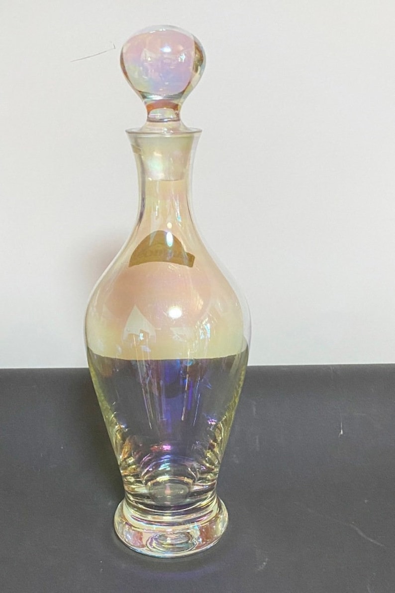May include: A clear glass decanter with an iridescent finish. The decanter has a round, bulbous body and a wide, round base. The top of the decanter has a round, iridescent stopper.