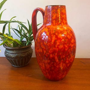 May include: A large, red and orange ceramic vase with a mottled glaze. The vase has a wide, round body and a single handle.