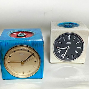 Puede incluir: Dos relojes de cerámica con forma de cubo. Uno es azul con esfera dorada y parte superior roja y turquesa. El otro es blanquecino con esfera negra. Ambos tienen la marca "Diehl electronic".