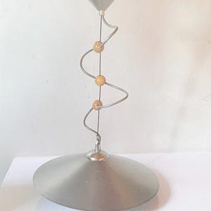 Könnte beinhalten: Eine silberne Pendelleuchte mit einem Spiraldesign und drei Holzperlen. Die Leuchte hat einen breiten, flachen, silbernen Schirm.