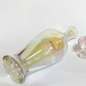 May include: A clear glass bottle with an iridescent finish and a matching glass stopper. The bottle has a round body and a long neck.