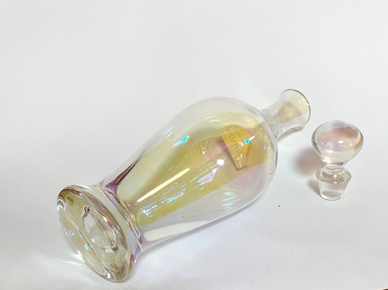 May include: A clear glass bottle with an iridescent finish and a matching glass stopper. The bottle has a round body and a long neck.