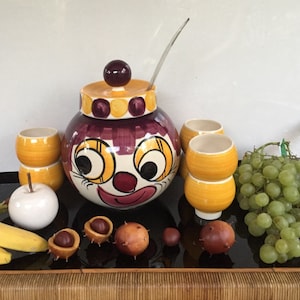 Puede incluir: Un tazón de ponche de cerámica con forma de cara de payaso con detalles amarillos, morados y blancos. El tazón tiene una tapa y una cuchara de vidrio transparente. Hay seis pequeñas tazas de cerámica amarillas y blancas, un racimo de uvas verdes y una manzana de cerámica blanca.