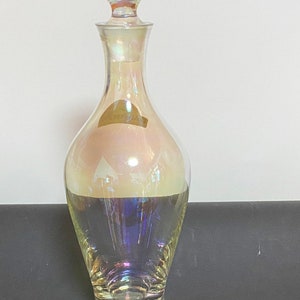 May include: A clear glass decanter with an iridescent finish. The decanter has a round, bulbous body and a tall, slender neck. The stopper is also round and iridescent.