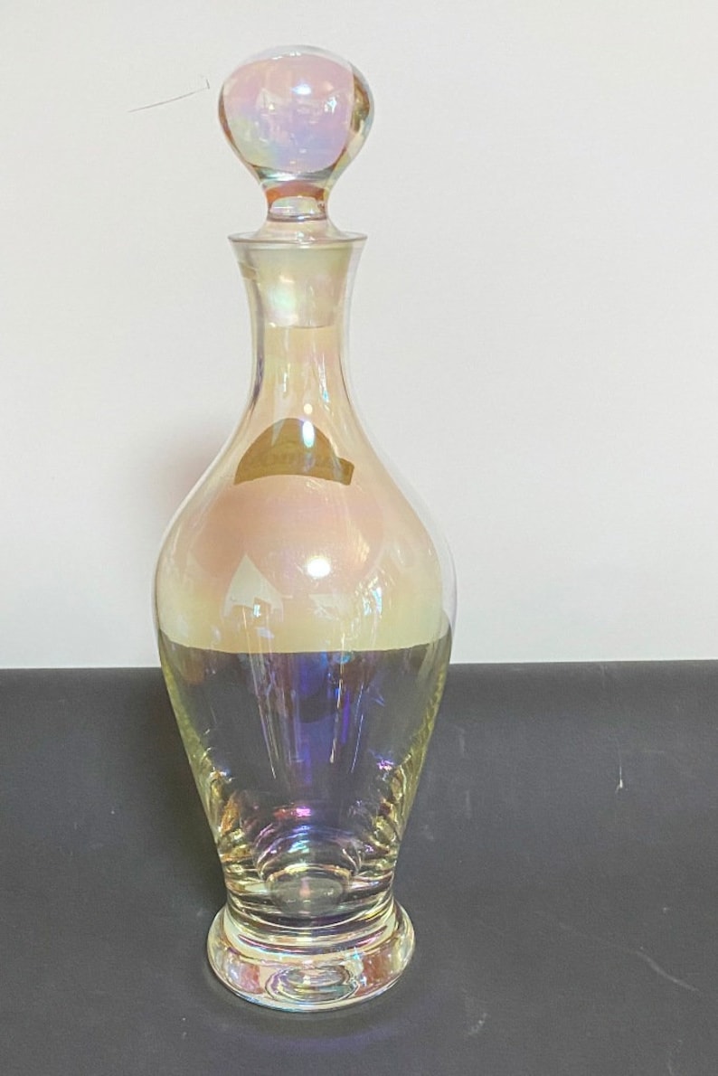 May include: A clear glass decanter with an iridescent finish. The decanter has a round, bulbous body and a tall, slender neck. The stopper is also round and iridescent.
