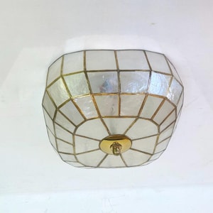 May include: A ceiling light fixture with a geometric design. The light has a white, iridescent surface with gold-coloured metal accents. The fixture is mounted on a white ceiling and has a brass-coloured centre.