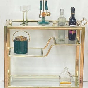 May include: A gold-toned bar cart with glass shelves and a handle. The cart holds a variety of barware, including wine glasses, bottles, a green ice bucket, and a decorative bottle stopper. The cart has a classic, elegant design.