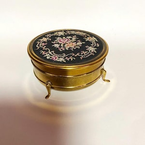 Könnte beinhalten: Eine runde, vintage Schmuckschatulle aus Messing mit einem schwarzen Stoffdeckel. Der Deckel hat ein florales Wandteppichdesign in Rosa, Weiß und Grün. Die Schatulle wird von drei kleinen, gebogenen Messingfüßen getragen, und die Seiten sind goldfarben poliert.