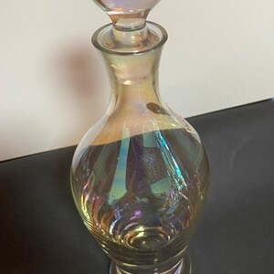 May include: A clear glass decanter with an iridescent finish. The decanter has a round, iridescent stopper and a wide base.