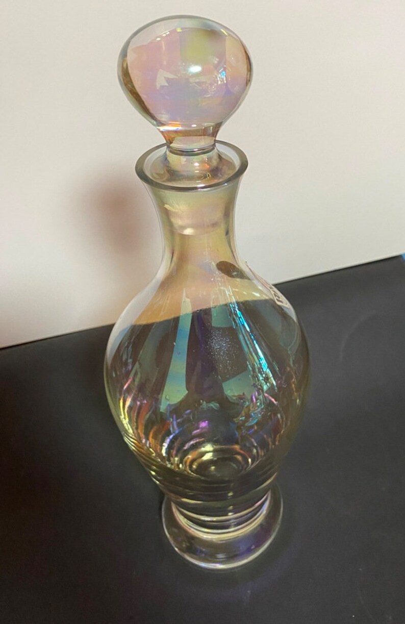 May include: A clear glass decanter with an iridescent finish. The decanter has a round, iridescent stopper and a wide base.